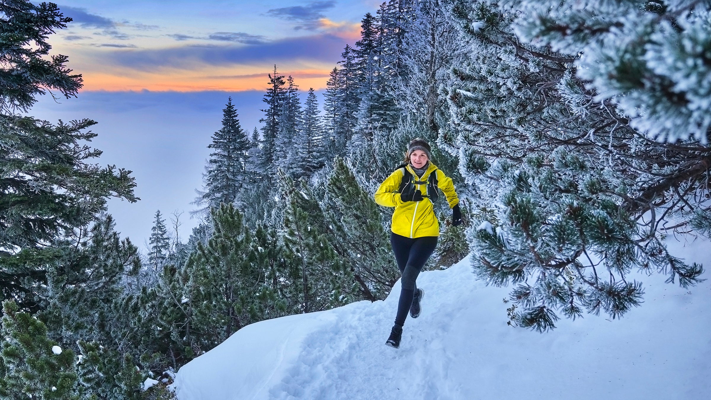 Woman in a yellow shell jacket hiking a snowy forest trail at dusk