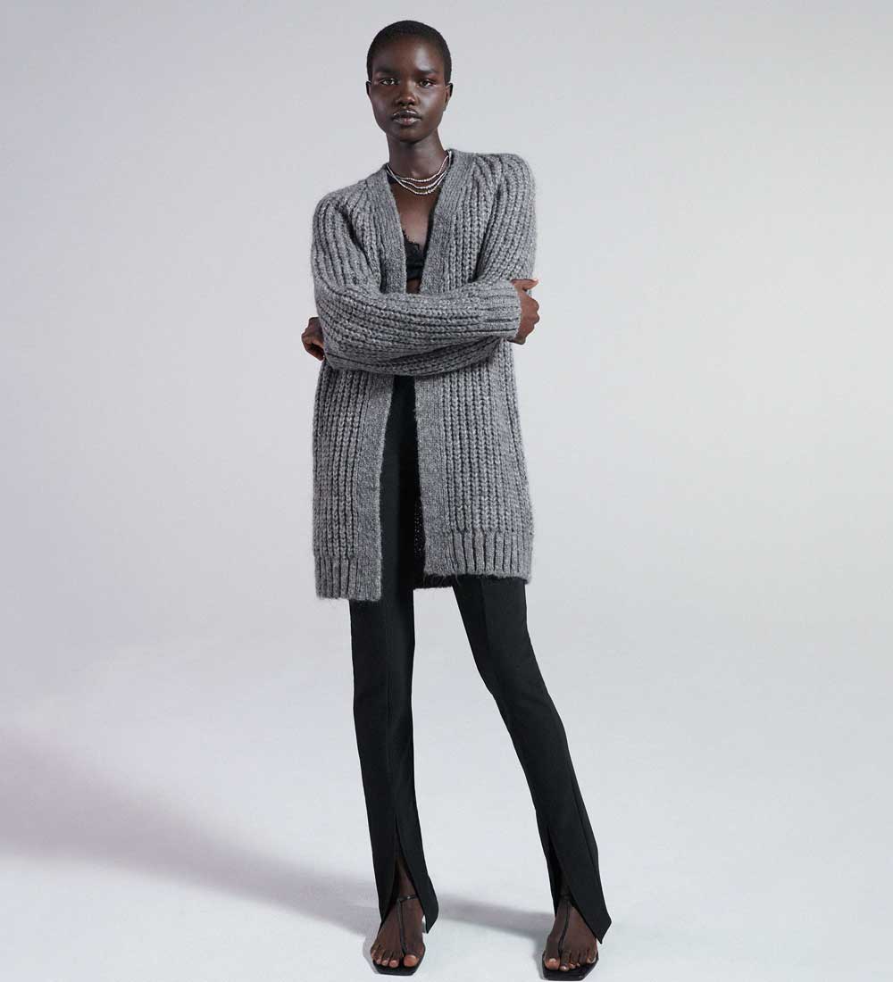 Woman wearing a long chunky grey knit cardigan open over a black top and black split-hem trousers with strappy heels