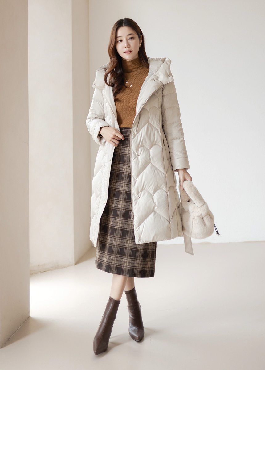 A woman wearing a long black puffer coat over a white pleated dress with a plaid scarf